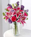 
Bright hot pink roses, fragrant stargazer lilies, beautiful purple lisianthus and larkspur are arranged with silver dollar eucalyptus in an urn-shaped glass vase...
			